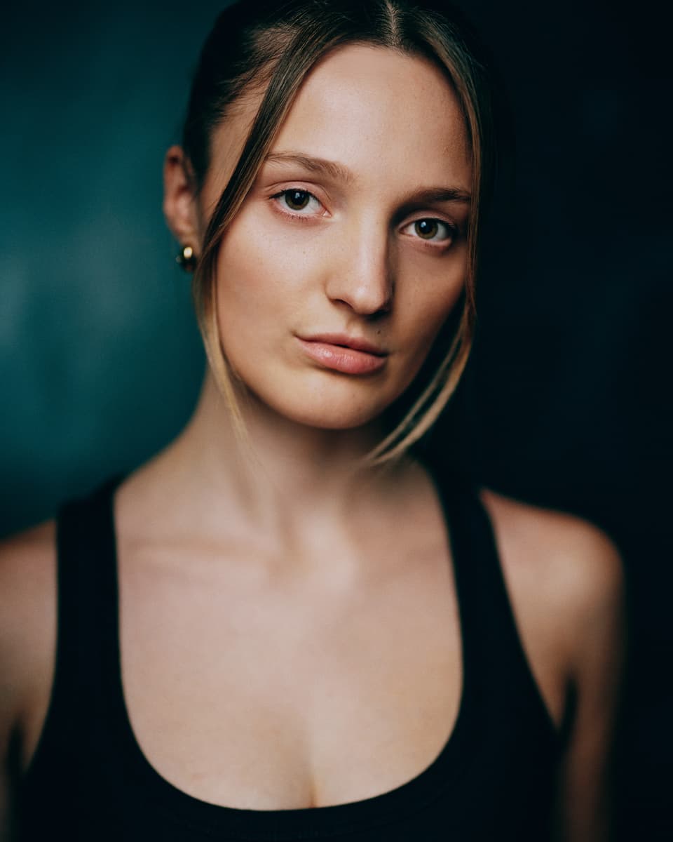 Toronto editorial actress headshot of a young woman with blonde hair swept back and gold hoop earrings, wearing a black tank top, with a calm and striking gaze against a dark teal background