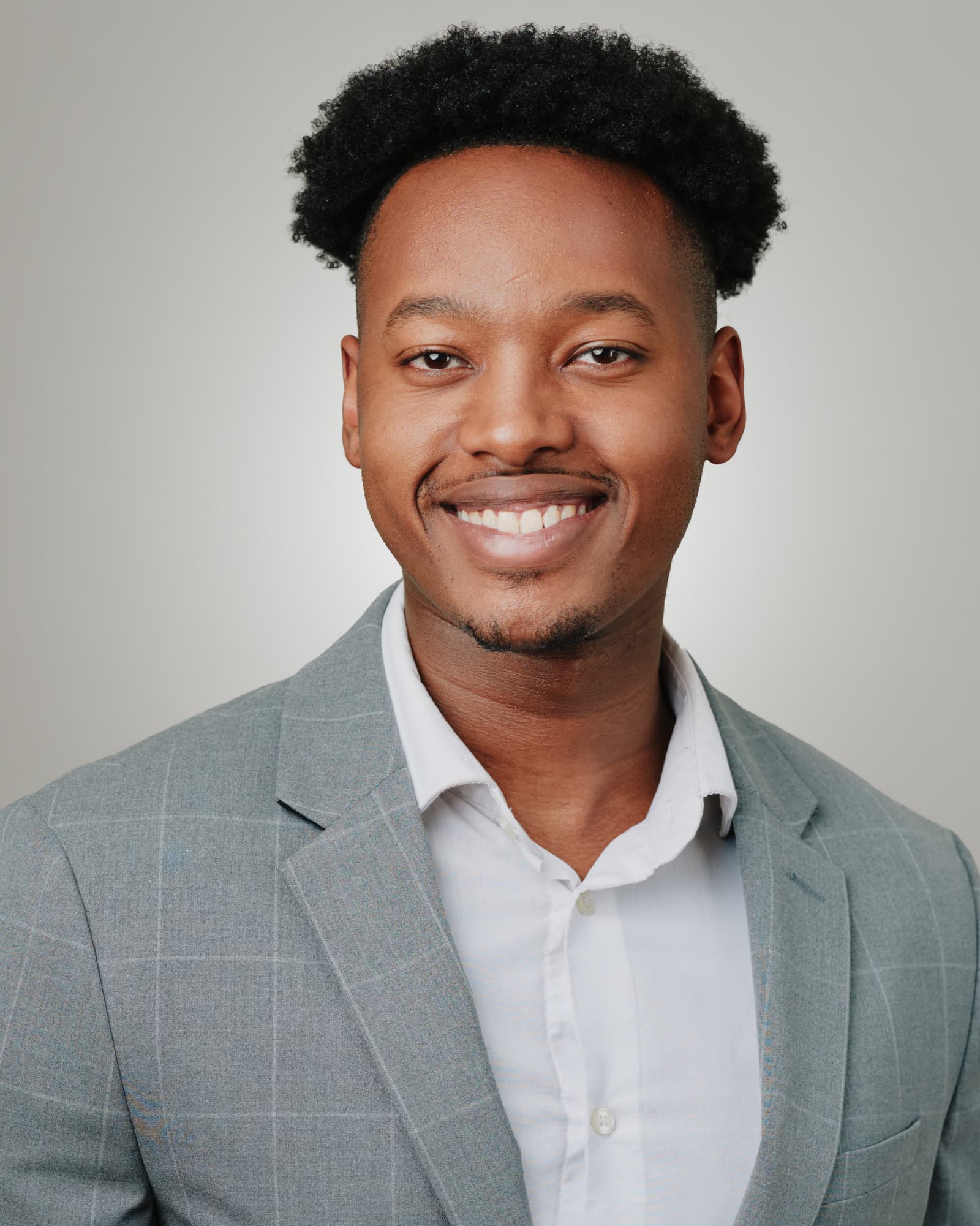 Toronto LinkedIn headshot of a young man with natural coily hair, wearing a light blue-grey plaid blazer over a white shirt, smiling broadly and confidently against a soft light grey studio background