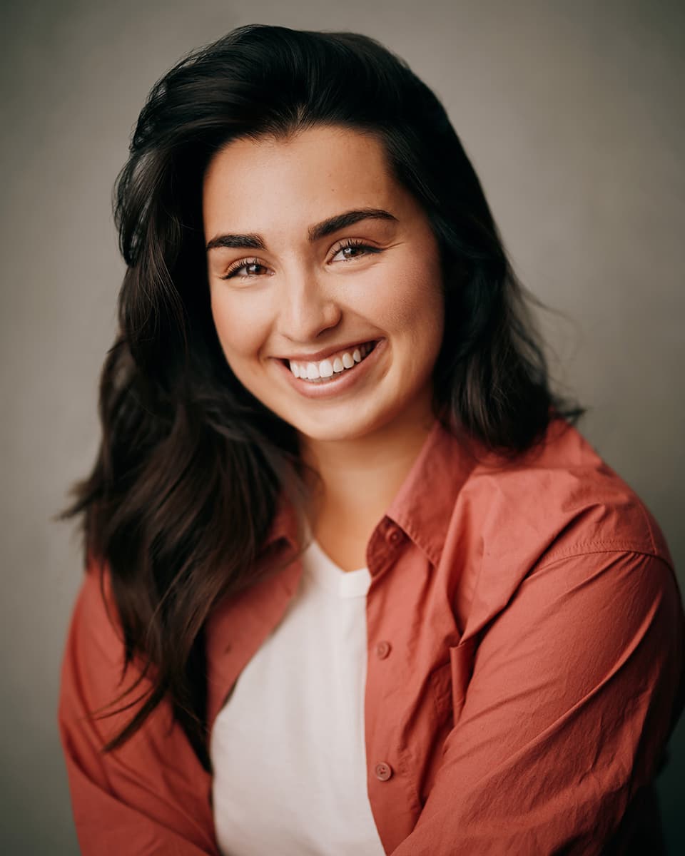 Toronto performing artist headshot of a young woman with long dark wavy hair in a terracotta button-up shirt, smiling warmly against a soft grey studio background